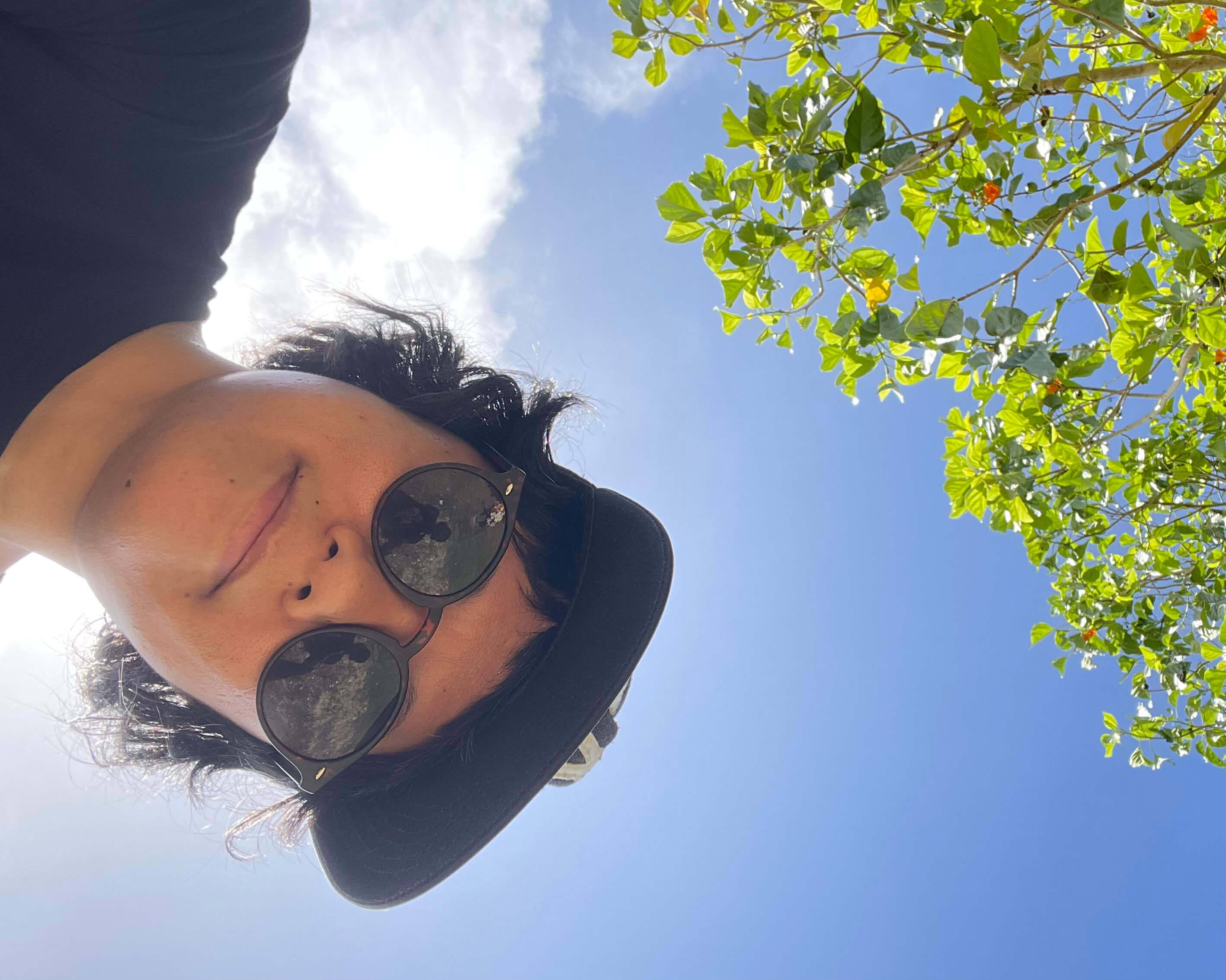 A brown nonbinary person wearing a black t-shirt, sunglasses, and baseball cap looking down at the camera and backlit by sunlight, a cloud, some tree branches, and the sky.